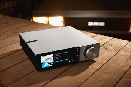 Schwarzer Audio-Video-Verstärker mit Dolby Atmos von vorne gesehen, LCD-Bildschirm zeigt "It's Alive!" an, auf einem Holztisch platziert
