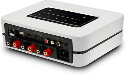 Integrovaný stereo hi-fi zesilovač BlueSound Powernode 2025 (N331) v bílé barvě, bílý obdélníkový plastový kryt s matným povrchem, černý dotykový panel zapuštěný na horní straně a kompletní zadní konektory (červené/černé svorky pro reproduktory, HDMI, USB, Ethernet, RCA), pohled zezadu ze tří čtvrtin
