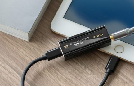 Pieni suorakulmainen musta digitaalinen-analoginen muunnin, ylhäältä ja viistosta kuvattuna, lasinen etupaneeli, jossa OLED-näyttö näyttää äänitietoja, ja jonka sivuilla on USB-C-portti sekä 3,5 mm kuulokelähtö, molemmat liitettyinä mustilla kaapeleilla