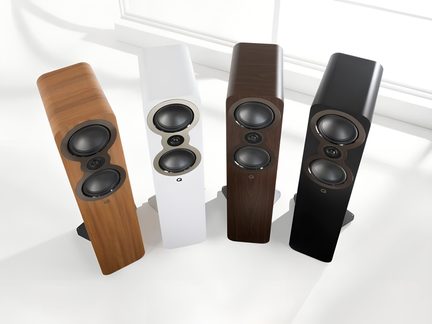 Four floor-standing speakers in light wood, matte white, dark wood, and satin black, seen from a three-quarter overhead angle, each featuring a smooth cabinet, two large circular drivers, and a central tweeter set in a sculpted figure-eight baffle