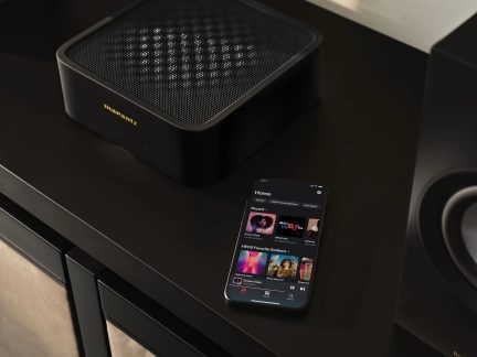 Svart forsterker med perforert grill, smarttelefon som viser en musikkapplikasjon, delvis synlig svart høyttaler