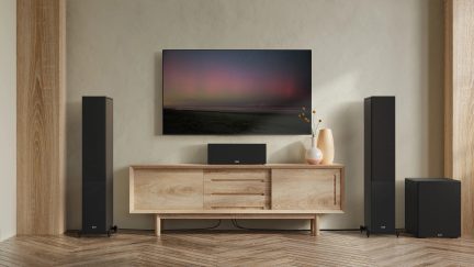 Lyst træmøbel med 3 skuffer og 2 låger, vægmonteret fjernsyn, 2 sorte gulvhøjttalere, subwoofer, beige vase