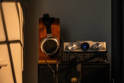 Căști STAX negre și argintii pe un suport, lângă un amplificator hi-fi cu două butoane rotative și ecran albastru