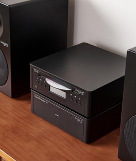 Zwarte metalen cd-speler op bijpassende versterker, met zichtbaar scherm, knoppen en sleuf, tussen twee luidsprekers, driekwartaanzicht van voren