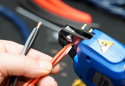Detailní záběr, šikmý pohled na ruku držící dva elektrické kabely, červený a šedý, s viditelnými měděnými vlákny, umístěné před černými čelistmi modrého kovového a plastového lisovacího kleště.