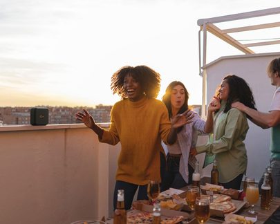 Quattro donne sorridenti su un balcone, che discutono attorno a un tavolo con cibo e bevande al tramonto