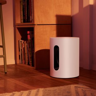 Mat beyaz, silindirik ve dikey Sonos Sub Mini subwoofer, parke zemin üzerinde, kitaplarla dolu bir kitaplığın yanında
