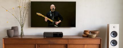 Ecran plat mural negru cu imaginea unui chitarist, bufet din lemn, player audio negru, difuzor coloană alb