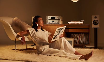 Forsiden viser en kvinne i hvitt på en myk teppe, vinylplateomslag, møbel i tre med hvit platespiller og sølvfarget forsterker