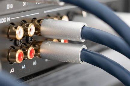 Tæt billede i skrå vinkel af to lydkabler med mørkeblå kappe og cylindrisk børstealuminiumsbeslag indsat i gyldne metal-RCA-stik med røde og hvide ringe på bagsiden af et sort hi-fi-apparat.
