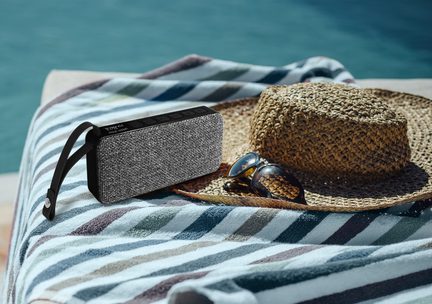 Enceinte portable rectangulaire grise en tissu, dessus noir à boutons, sangle latérale, vue de trois-quarts sur serviette rayée