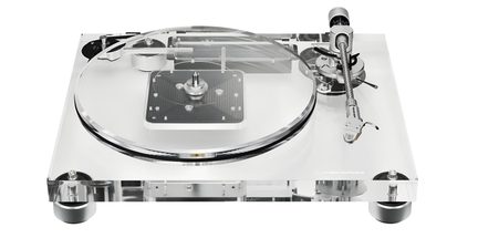 Transparent akryl-pladespiller med klar, cirkulær pladetallerken, sølvfarvet metal-tonearm til højre og sorte cylindriske fødder, set let oppefra forfra