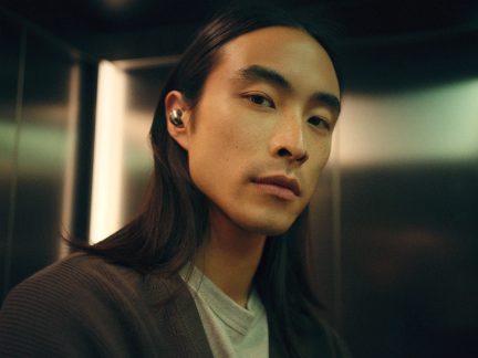 Auricolare wireless grigio indossato nell’orecchio sinistro, ritratto ravvicinato di un uomo con lunghi capelli neri