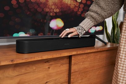 Barra audio Sonos Beam (Gen 2) nera opaca appoggiata su un mobile in legno, mani femminili che toccano il dispositivo, sfondo colorato sfocato