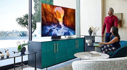 Televizor Samsung QE75Q90R zobrazující přírodní scénu v moderním obývacím pokoji s tyrkysovou pohovkou, rostlinami a velkými okny