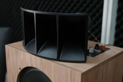 Haut-parleur en bois clair avec pavillon noir, boutons et connecteurs visibles sur le dessus, angle de vue légèrement en plongée