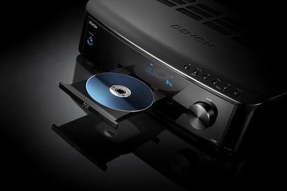 Czarny odtwarzacz Blu-ray Denon S-5BD, widok z przodu w perspektywie, z otwartą tacą zawierającą niebieską płytę, na ciemnoszarym tle