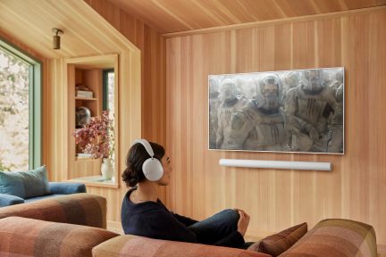 Persona seduta su un divano con plaid, cuffie bianche, schermo a parete con film, soundbar bianca, parete in legno, finestra a sinistra.