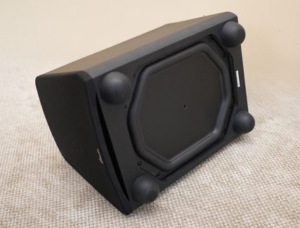 Alto-falante preto inclinado, visto de baixo, base octogonal, 4 pés esféricos pretos, superfície apoiada em um tecido bege