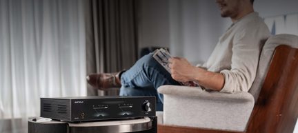 Černé hi-fi audio zařízení z kovu, pohled ze tří čtvrtin, tři VU metry, kolečko vpravo, konektor na čelním panelu, boční větrací otvory