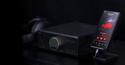 Juodo metalo garso stiprintuvo, matomo iš trijų ketvirčių priekio, su didele auksine rankenėle, apjuosta mėlyna šviesa, pastatyto ant tamsaus stalo, šalia kurio gale yra juodos ausinės ir įstrižai pastatytas stačiakampis juodas muzikos grotuvas, vaizdas.