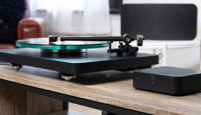 Μαύρο ματ δικτυακό ηχοσύστημα BlueSound HUB με ενσωματωμένη πικάπ, φωτογραφημένο μπροστά σε ξύλινο τραπέζι