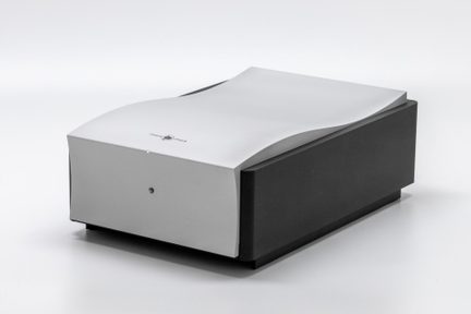 Caja electrónica rectangular de metal mate, laterales negros, parte superior de aluminio plateado con superficie ondulada, indicador LED redondo centrado en el panel frontal, vista en tres cuartos desde el frente izquierdo