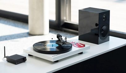 Bílý gramofon Pro-Ject Debut III Rolling Stones zepředu, s gramofonem a černým krychlovým reproduktorem