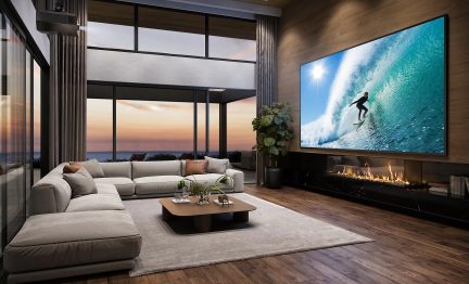 Moderne stue set fra hjørnet, lysegrå stofhjørnesofa på brun parket, stor vinduesfacade med solnedgang til venstre, kæmpe vægmonteret tv viser en bølge over en lang sort lineær pejs, træ-metal sofabord og lyst tæppe i midten