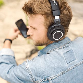 Černé sluchátko s kulatým stříbrným logem, nasazené na hlavě se světlými vlasy, světle modrá džínová košile, držení černého smartphonu