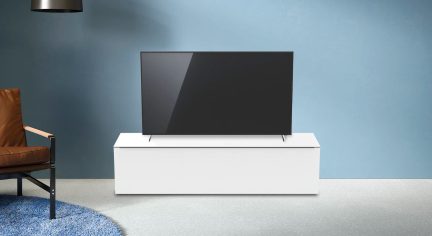 Černý vypnutý televizor položený na bílém obdélníkovém nábytku v místnosti se světle modrou stěnou, pohled zepředu