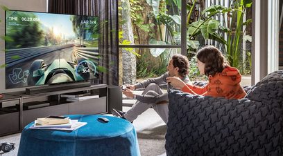 Billedet viser to børn, der sidder på en grå sofa og spiller et bilracerspil på en stor fladskærms-tv.