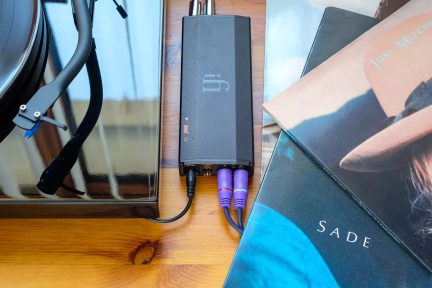 Sort pladespiller med synlig tonearm, sort rektangulær forstærker forbundet med lilla kabler, stablede albumcovers