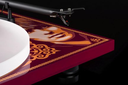 Rektangulær rød plade med orange mønstre og stiliseret ansigt, sort tonearm, hvid cirkulær plade, sort baggrund