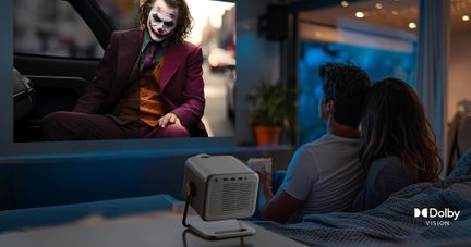Šedý plastový krychlový projektor světlejšího odstínu s bočními větracími mřížkami, umístěný na stolním stojanu a zobrazený z tříčtvrtečního pohledu zepředu, promítající v pozadí filmovou scénu před posedávajícím párem.