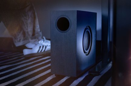 Subwoofer til hjemmebiograf i sort træ, sidehøjttaler og frontventilation, placeret på stribet tæppe i moderne interiør