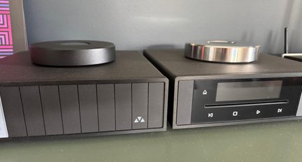 Pohled zepředu na dva obdélníkové hi-fi přehrávače z černého kovu umístěné vedle sebe, každý s velkým kovovým diskem nahoře – matně černým vlevo a leštěným stříbrným vpravo –, první s přední stranou s vertikálními drážkami a malým trojúhelníkovým logem, druhý s tmavou obrazovkou, horizontální štěrbinou a dotykovými ovládacími ikonami.