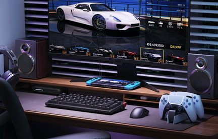 Escritorio de madera, vista frontal: pantalla con juego de carreras, altavoces metálicos, consola portátil azul, dos mandos blancos