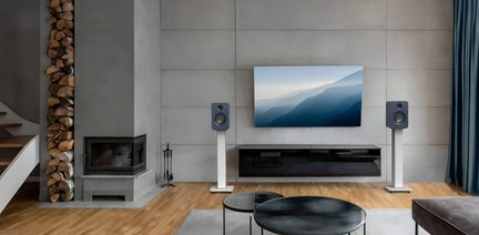 Frontansicht eines modernen Wohnzimmers, graue Betonwand mit zentriertem Flachbildfernseher, zwei kompakte dunkelblaue Lautsprecher auf weißen Metallständern an beiden Seiten, darunter eine schwebende, schwarz lackierte Konsole, heller Parkettboden und links ein Betonkamin mit vertikalem Holzlager.