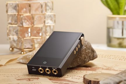 Dispositivo elettronico portatile rettangolare in alluminio nero, con schermo touch frontale lucido, tre porte audio dorate e connettore USB-C sul bordo inferiore, rotella del volume zigrinata e tre pulsanti circolari dorati sul lato destro, presentato in leggera prospettiva e appoggiato inclinato su una pietra