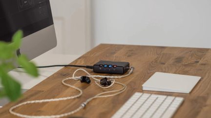 Lille, mat sort, bærbar lydkasse i metal set fra trekvart forfra, udstyret med farvede knapper på oversiden og forbundet til sorte in-ear-hovedtelefoner med sølvfarvet snoet kabel, placeret på et lyst træskrivebord