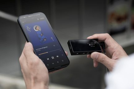 Player audio digital portabil negru cu ecran, ținut în mână lângă un smartphone pe care rulează un album de muzică