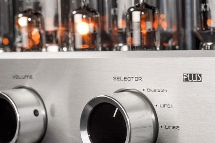 Mierne šikmý detailný záber na strieborný hliníkový čelný panel hi-fi zosilňovača s dvoma veľkými valcovými chrómovanými ovládačmi s čiernym stredom pre hlasitosť a volič, s vygravírovanými štítkami Bluetooth Line1 Line2 a rozmazanými žiariacimi elektrónkami v pozadí