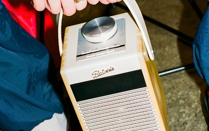 Radio portatile color crema e legno chiaro vista da tre quarti leggermente dall'alto, custodia rettangolare rivestita in pelle color crema con bordi in legno, grande ghiera metallica argentata sulla piastra superiore, maniglia arcuata abbinata, pannello frontale con fascia nera e griglia forata decorata con l'iscrizione « Roberts »