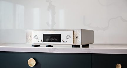 Player audio de rețea rectangular, jos, din metal argintiu, vedere din față, fațadă frontală texturată cu ecran central negru, două butoane circulare simetrice, buton rotativ mic, mufă pentru căști în dreapta și patru picioare negre