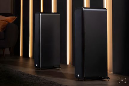 Two black floorstanding speakers in a matte wood cabinet with fabric grille, rounded edges, and a small white logo at the bottom, shown three-quarters from the front on spiked feet