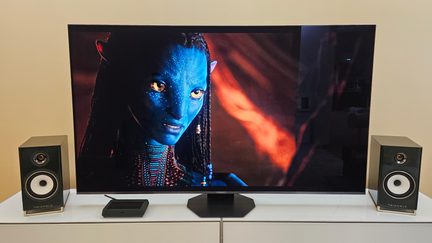 Televisore a grande schermo su mobile bianco, immagine Avatar, con due diffusori Triangle neri e box multimediale