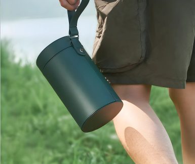 Cilindro verde scuro con manico, tenuto da una mano, sfondo di erba verde, gamba in pantaloncini marroni visibile