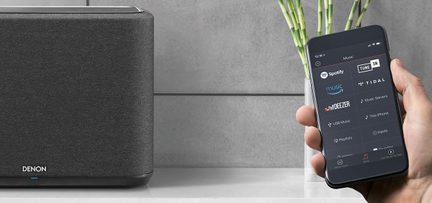 Rectangular black speaker and black smartphone displaying the Spotify app with green plants in the background