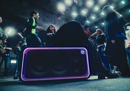 Svart bærbar høyttaler med grillfront og lilla lysende kant under en innendørs dansefest